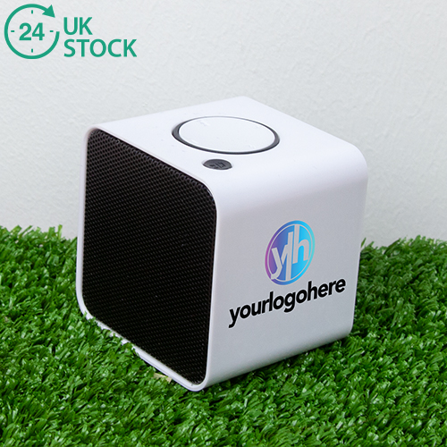 Cube bluetooth speaker printed speaker with 'your logo here' branded on. Sitting on a grass background with an icon that says 24 hours UK stock.